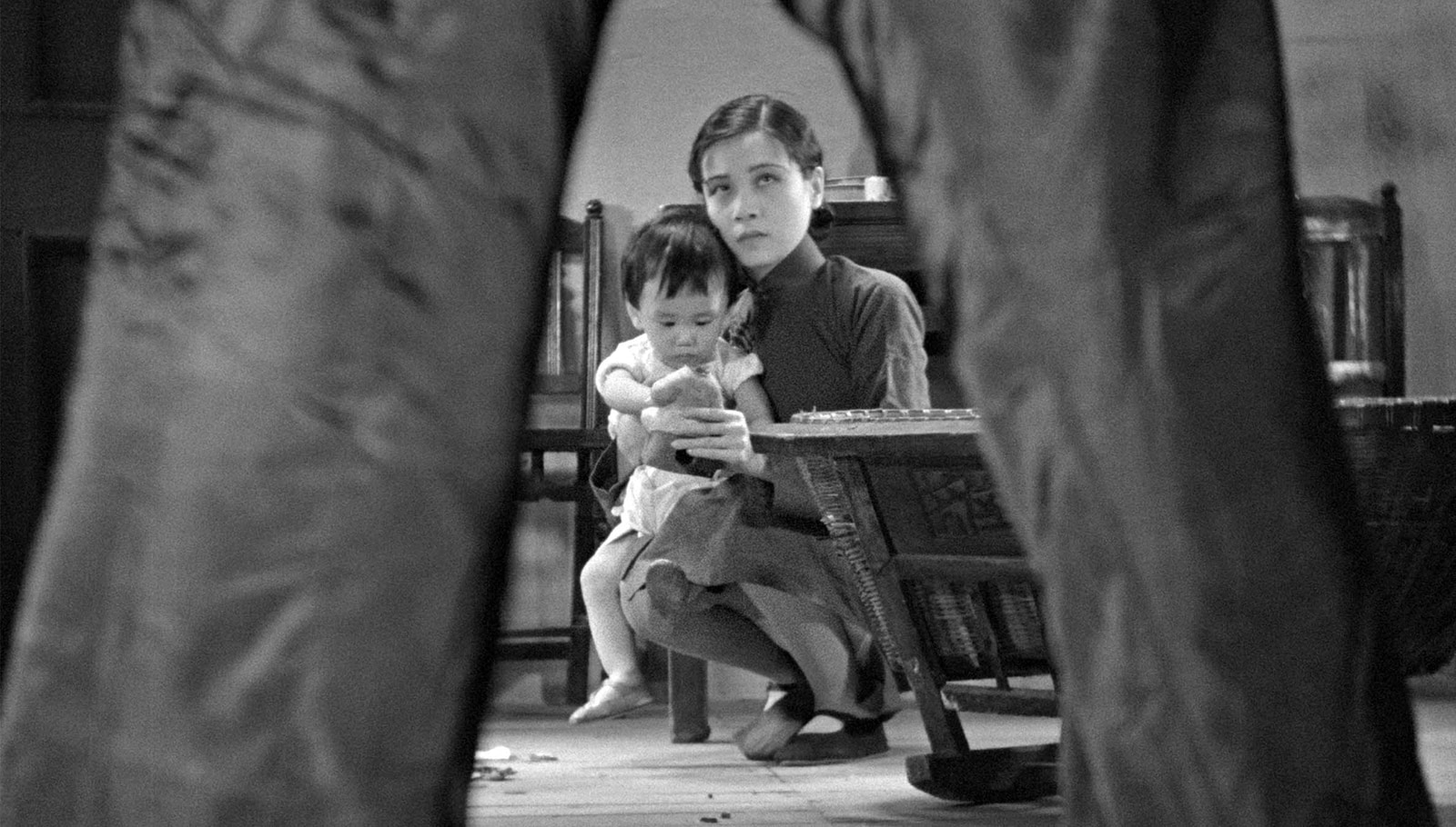 Black-and-white frame from The Goddess showing Ruan Lingyu seated with her infant son, seen through the legs of the Boss standing in the foreground.