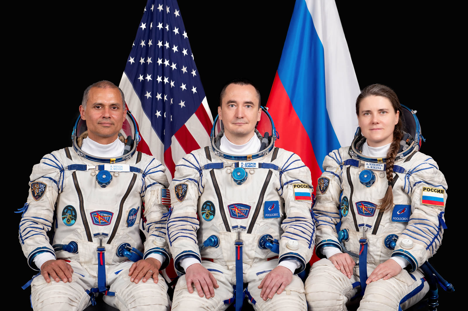 NASA 宇航员阿尼尔·梅农与 Roscosmos 宇航员彼得·杜布罗夫、安娜·基基娜身穿 Sokol 发射服，在美国和俄罗斯国旗前合影。
