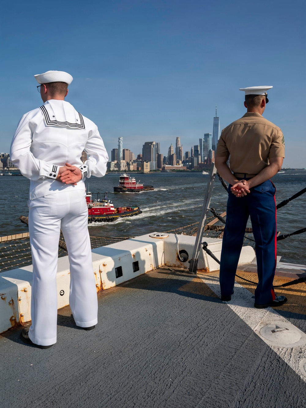 A sailor and a Marine stand on the deck of USS Arlington while the ship enters New York Harbor with the Manhattan skyline ahead.