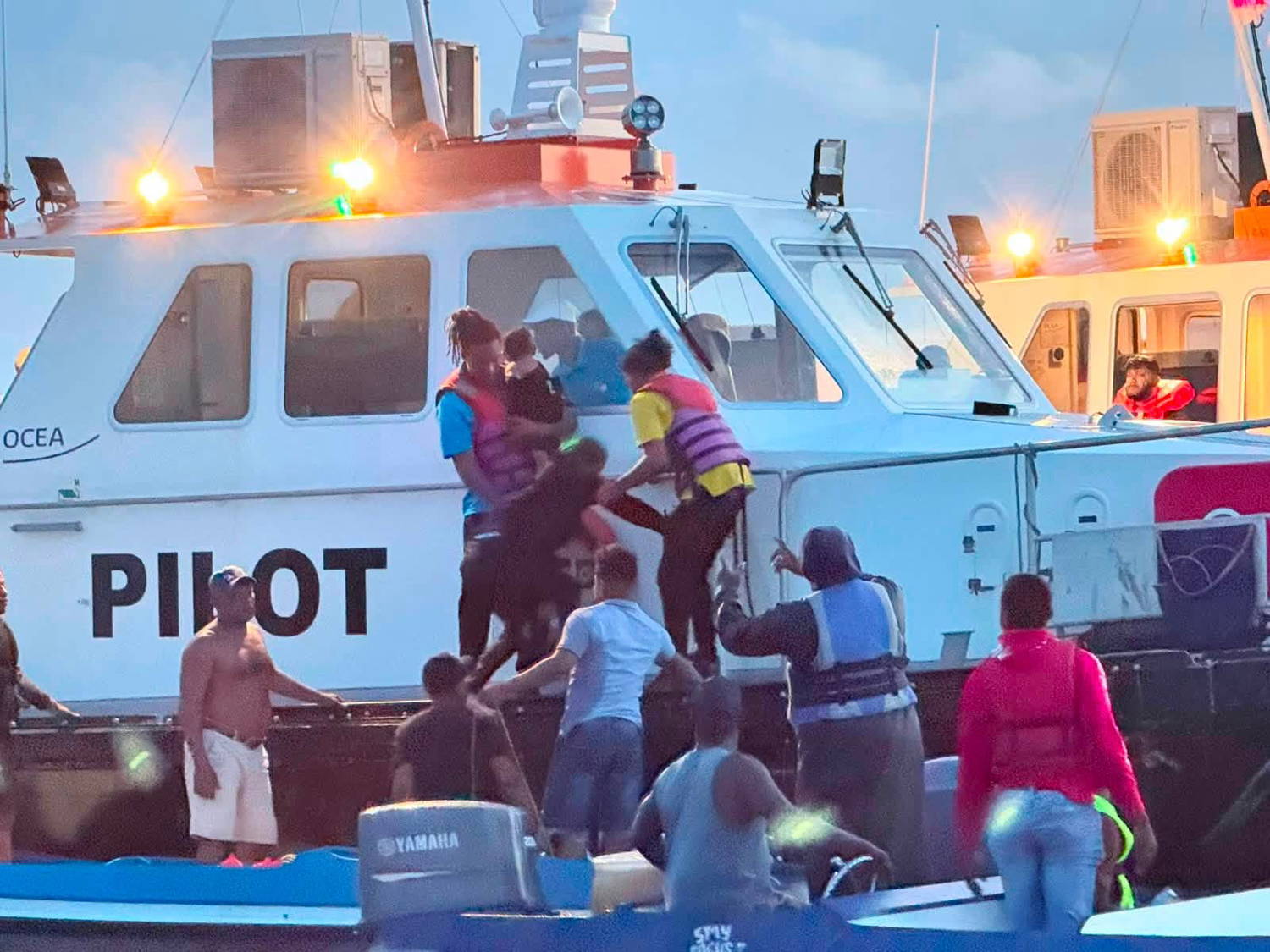 Rescuers in life jackets help adults and children transfer from a small boat onto a white pilot vessel after the MV Barima capsized off Guyana.