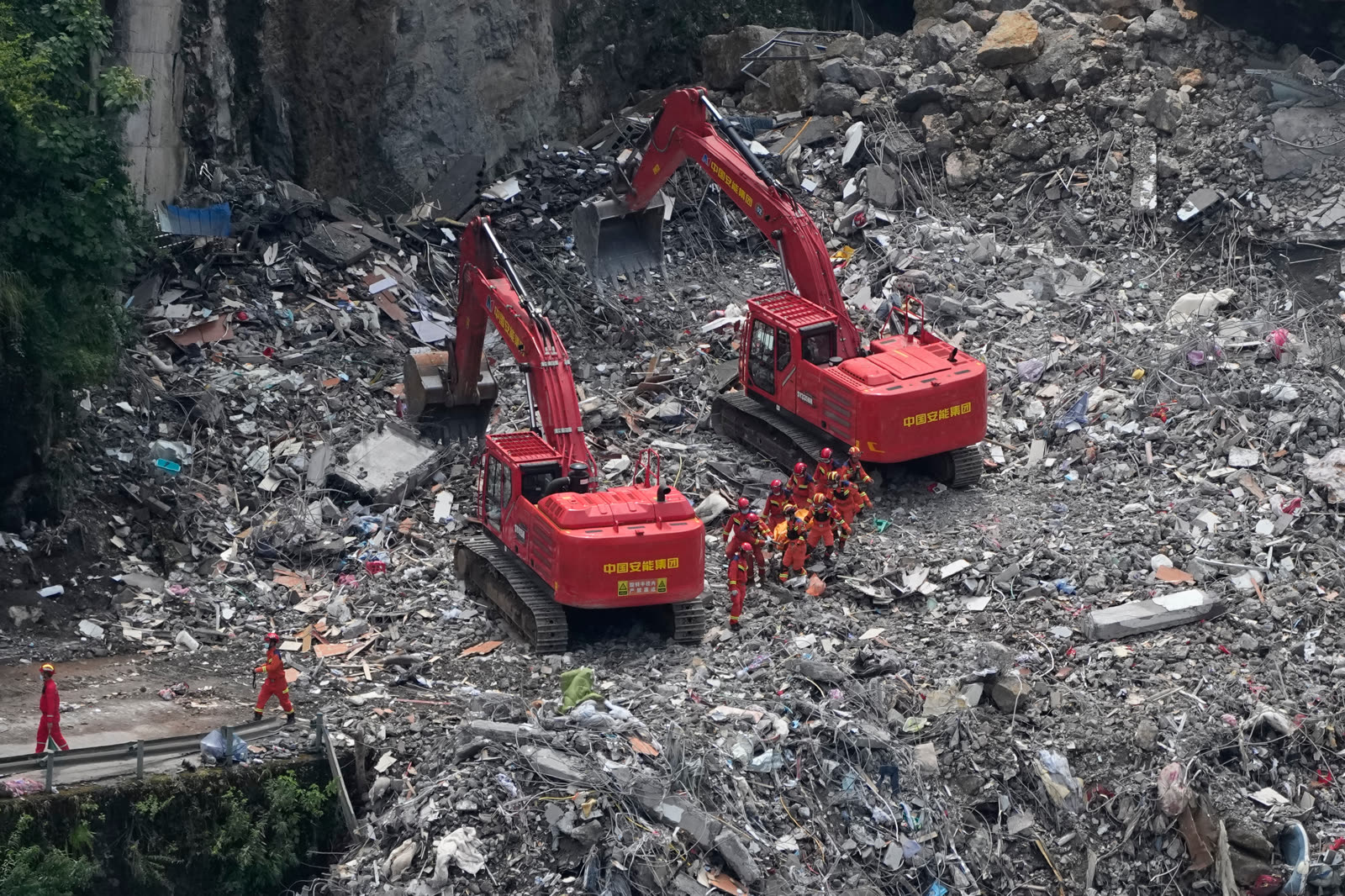 Rescue workers in orange and red uniforms move across the rock-strewn Pengshui landslide site as excavators work below the exposed slope.