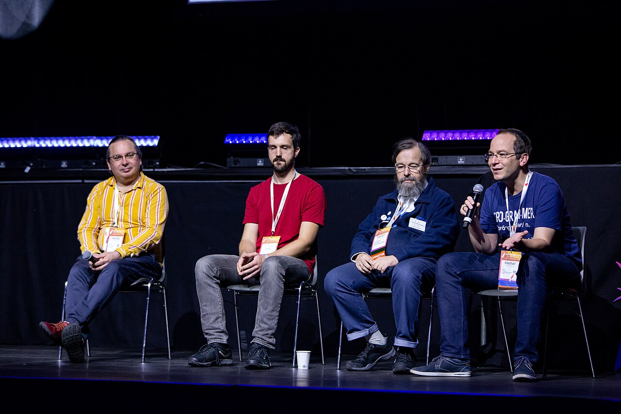 Four ApacheCon Europe panelists sit on a dark stage while one speaks into a handheld microphone.