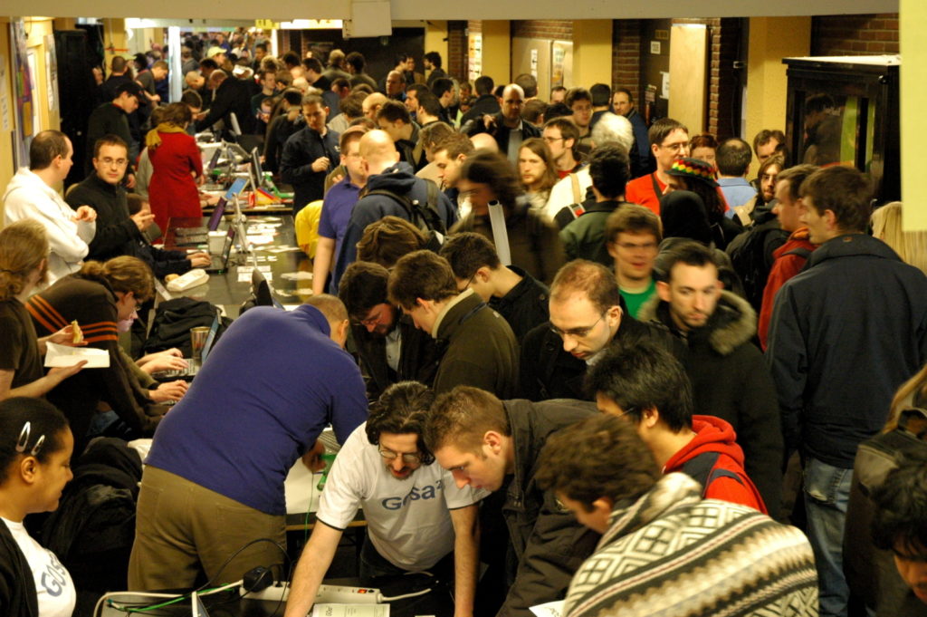 Crowded Fedora Project booth at FOSDEM with contributors and visitors gathered around laptops and tables.