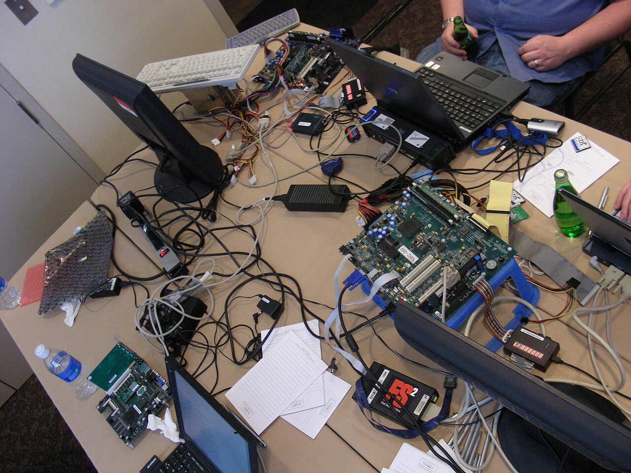在 coreboot 2008 年丹佛峰会的工作桌上，打开的笔记本电脑、裸露的主板、编程设备和线缆铺满桌面，两名贡献者的身影局部入镜。