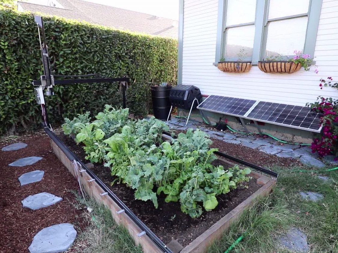 An early FarmBot Genesis gantry installed over a raised garden bed filled with leafy crops beside a house.