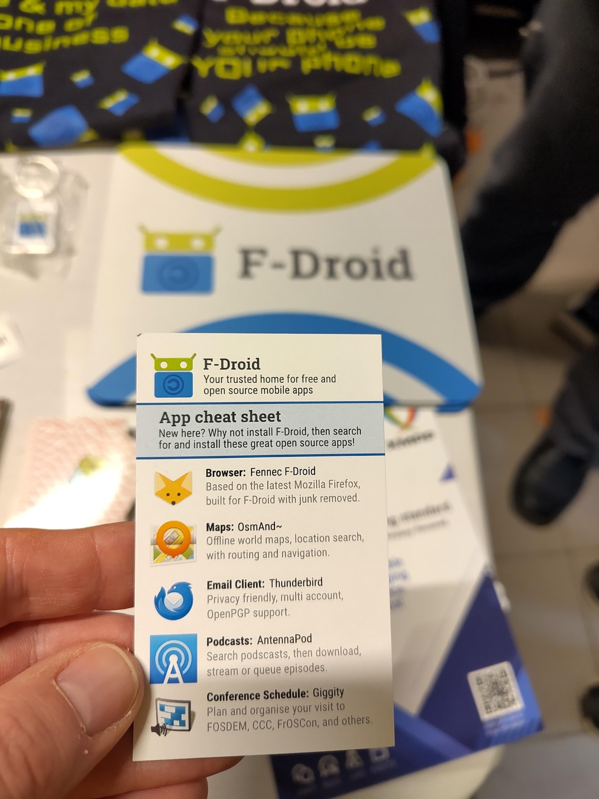 A hand holds an F-Droid app cheat sheet in front of the F-Droid stand at FOSDEM 2026.