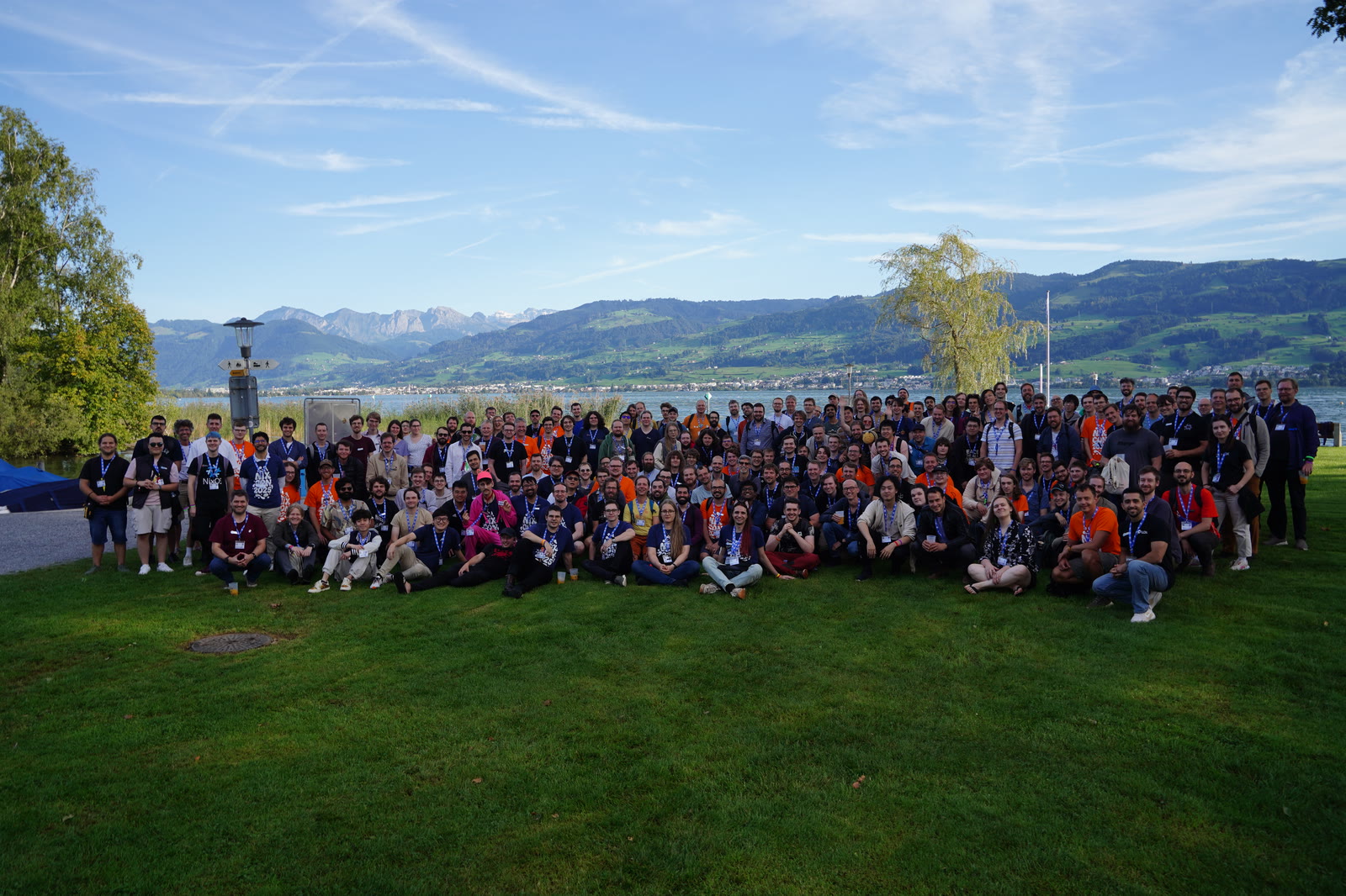 Hundreds of NixCon 2025 attendees standing together on a lawn beside a lake, with wooded hills and mountains behind them.