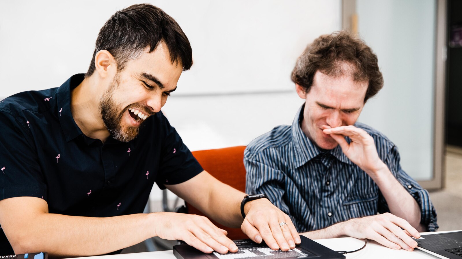 NV Access founders Jamie Teh and Michael Curran sit together at a table, smiling as they feel a prototype braille graphic display.