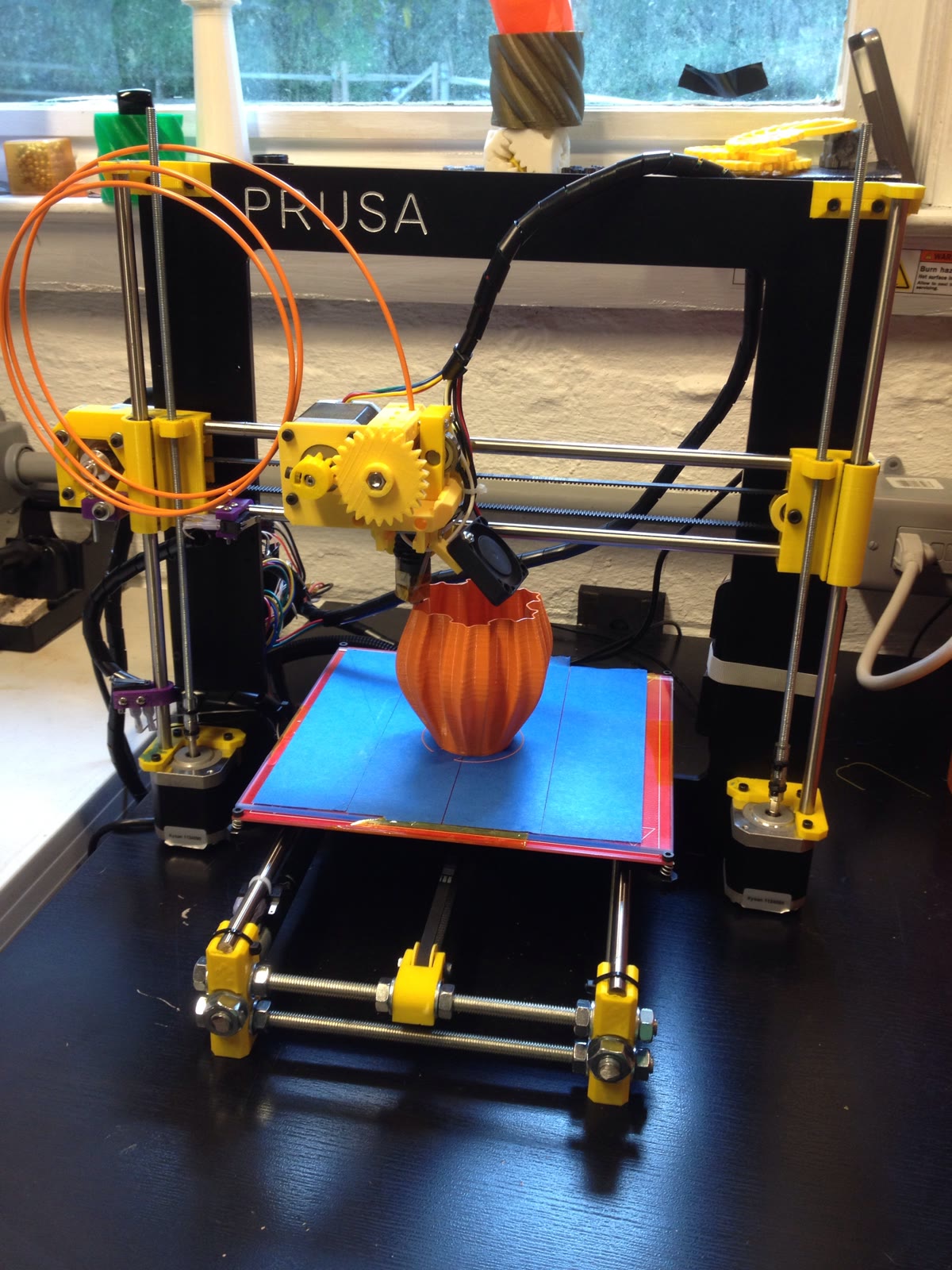 A black-and-yellow RepRap Prusa i3 printer laying orange filament into a vase on a blue print bed.