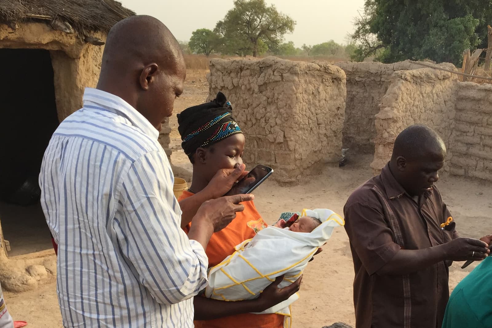 A field worker uses a smartphone while standing beside a woman holding a newborn in a rural compound in Mali.
