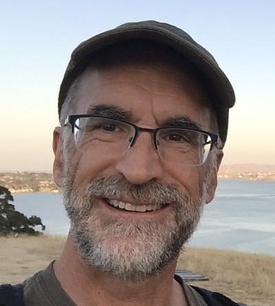 John MacFarlane 戴着眼镜和帽子，在水边户外微笑的照片。