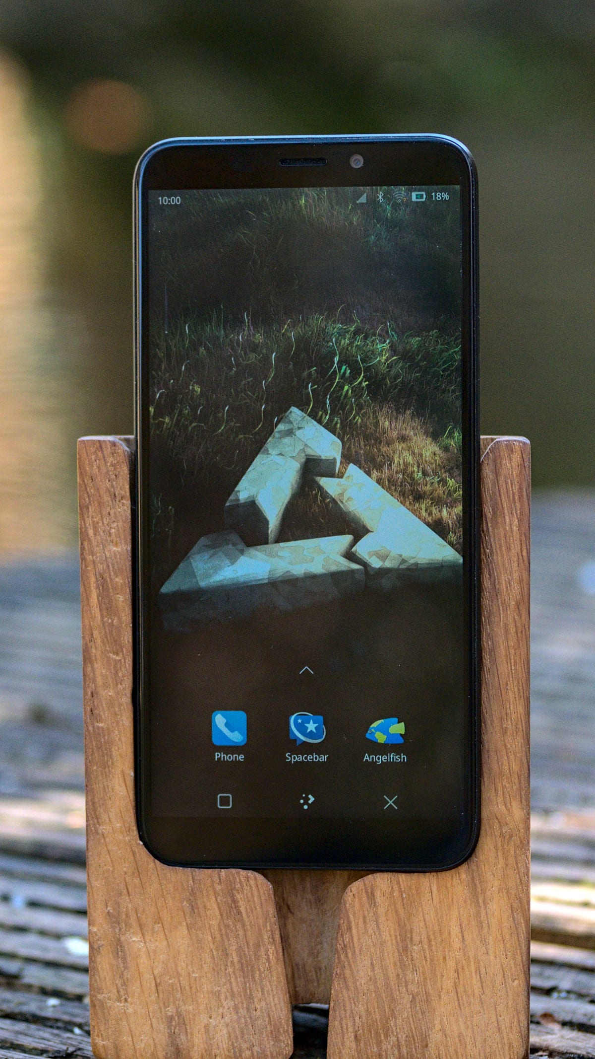 A PinePhone Pro displaying the Plasma Mobile home screen while resting in a wooden stand outdoors.