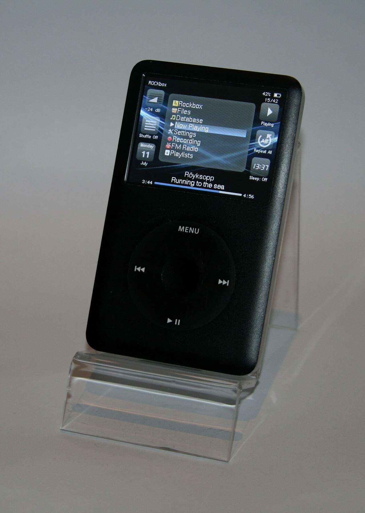 A black sixth-generation iPod classic on a clear stand, its screen showing the Rockbox main menu and playback controls.