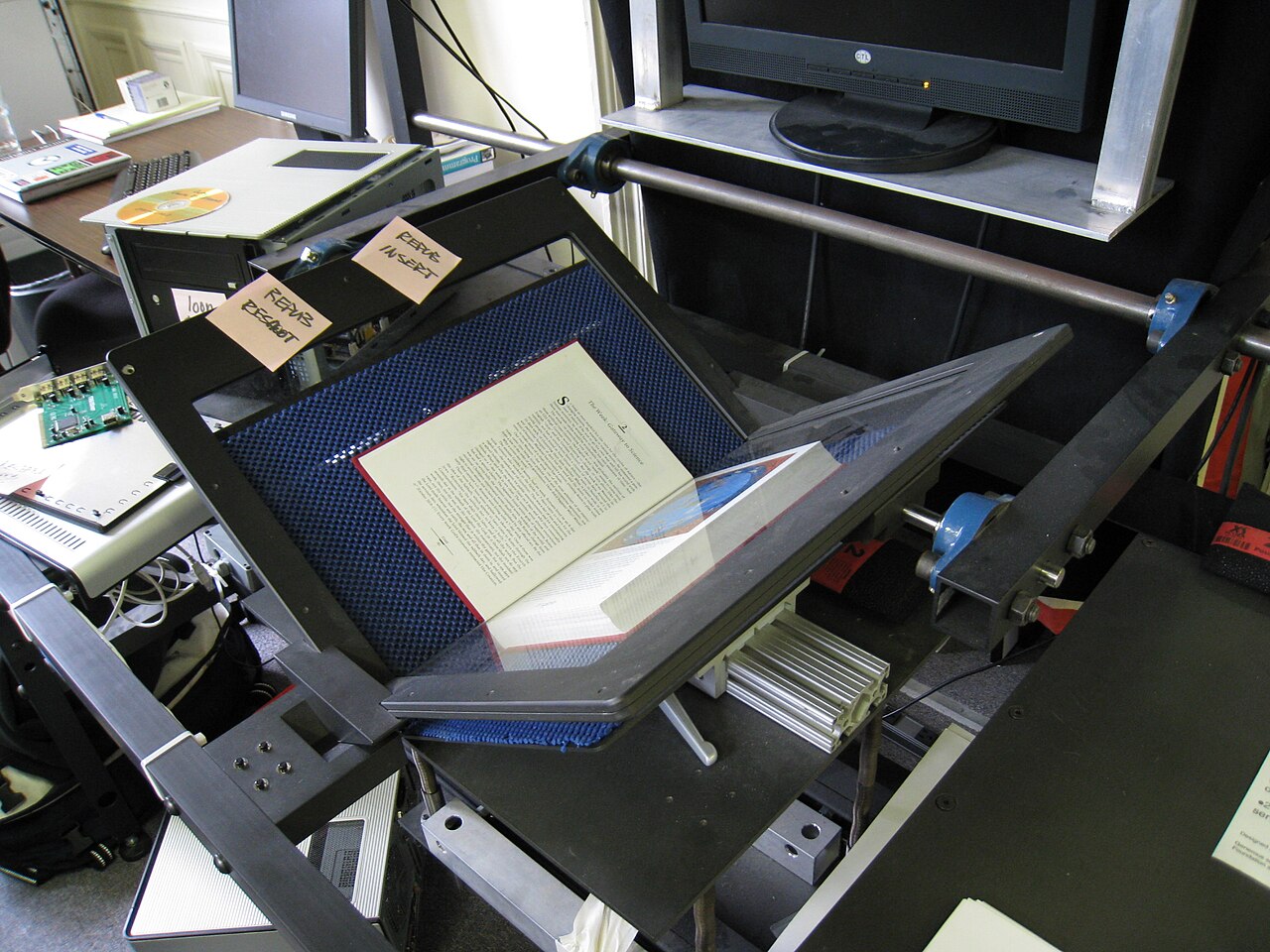 A book rests under a glass platen inside an Internet Archive scanning station, with cameras, a monitor, and metal scanner rails around it.