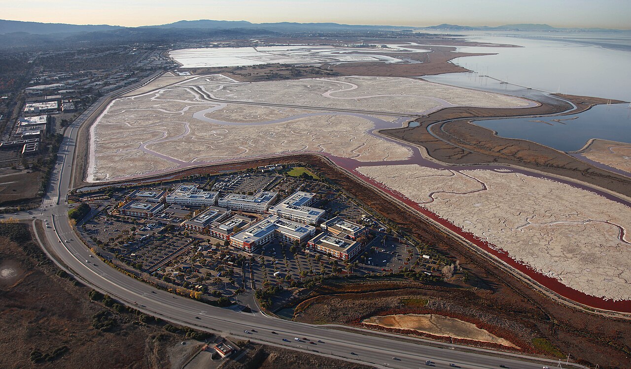 Facebook 位于 Menlo Park 的总部园区航拍照片，可见建筑、道路，以及远处的 San Francisco Bay。