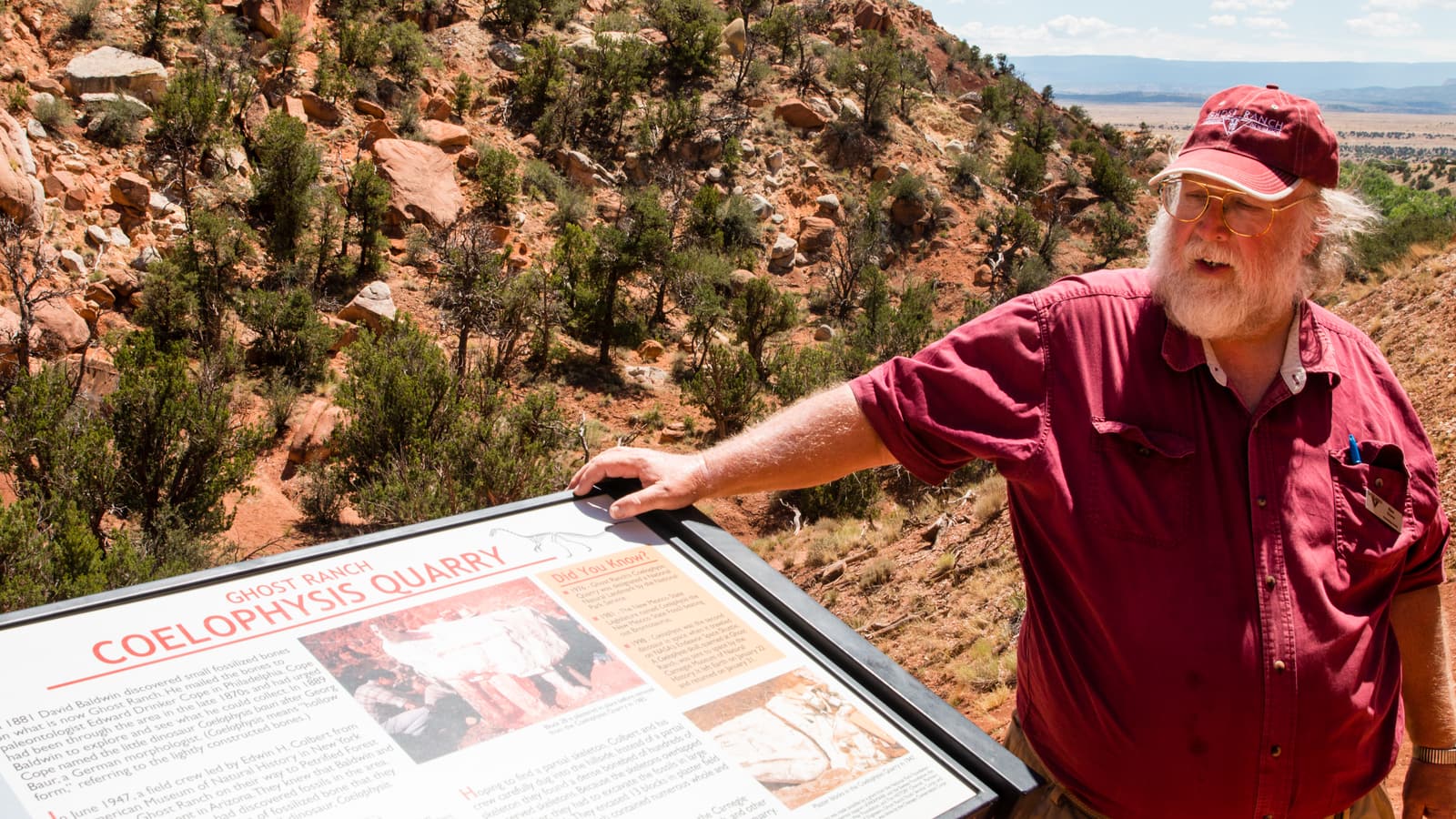 A white-bearded guide in a red shirt stands beside the Coelophysis Quarry sign at Ghost Ranch, with red Chinle Formation slopes behind him.