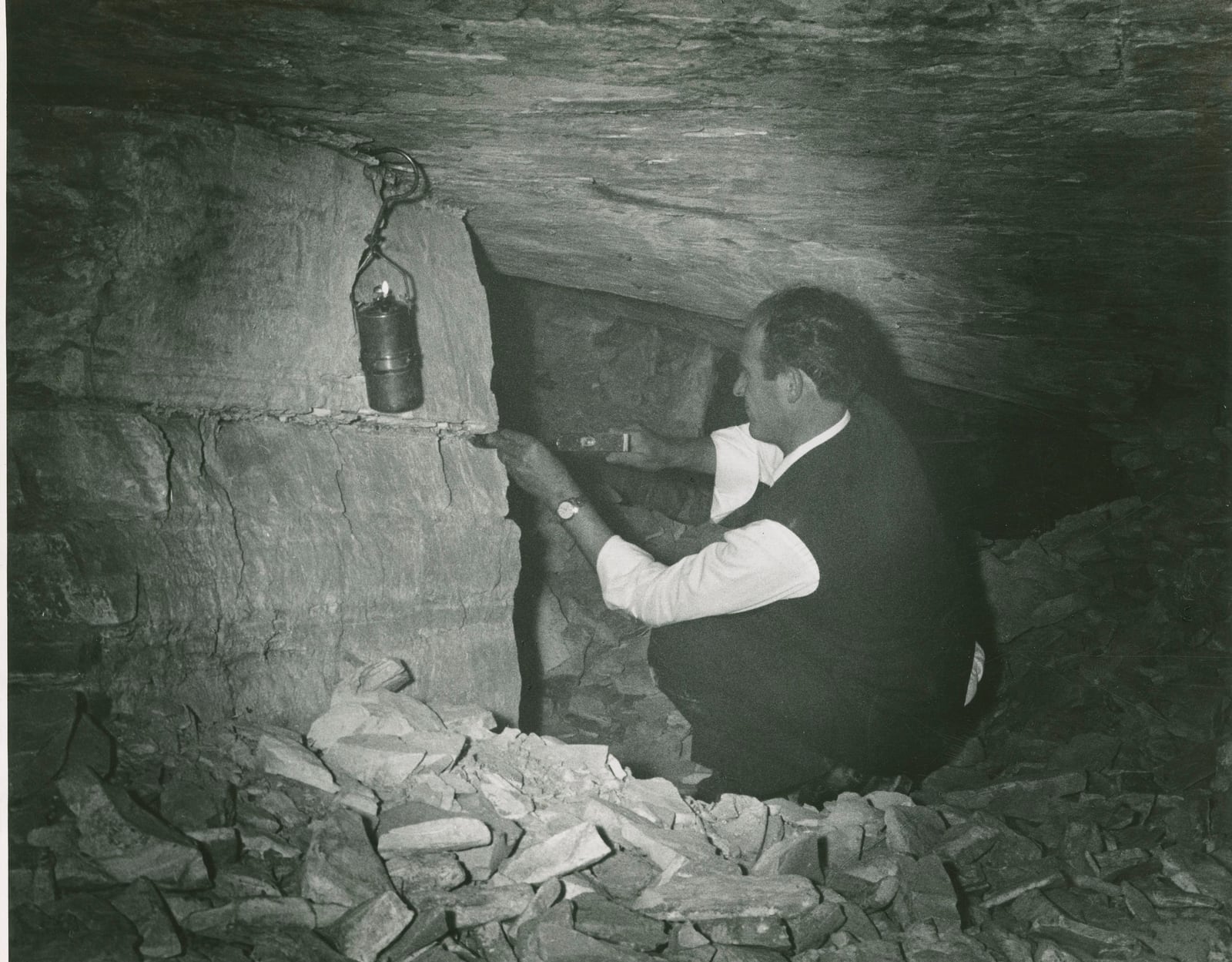 Black-and-white archival photograph of Massimiliano Cerato kneeling among stacked limestone inside the Pesciara quarry while extracting a fossil in 1964.