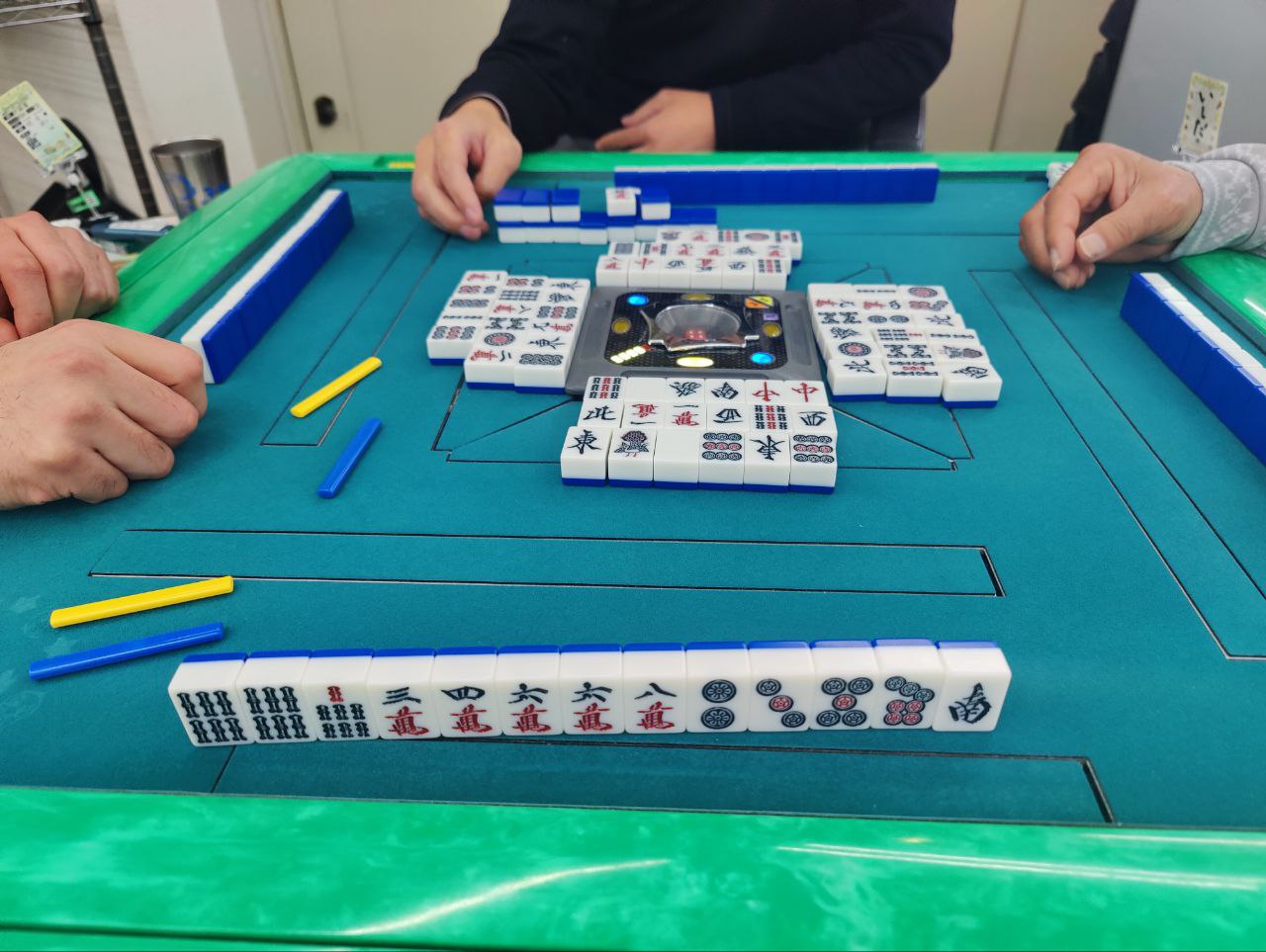 Four people playing Japanese riichi mahjong around an automatic table, with tile walls, discards, and scoring sticks visible.