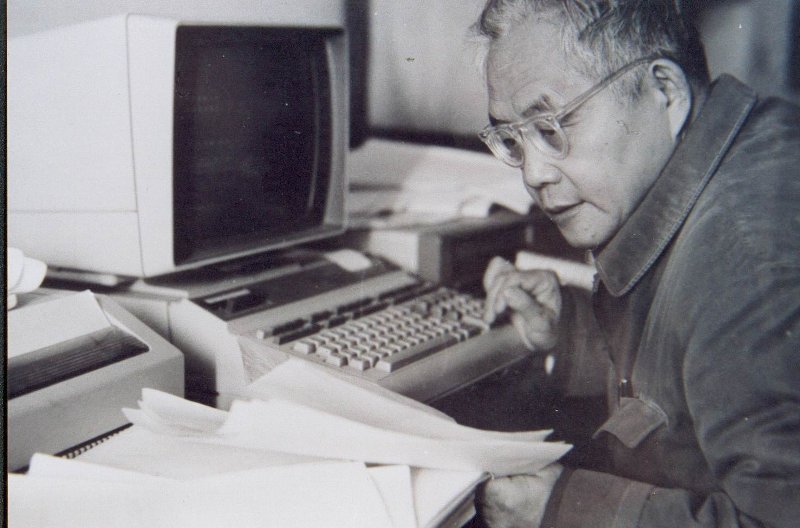 一张黑白档案照片：数学家吴文俊坐在个人计算机旁，正在阅读一沓论文。