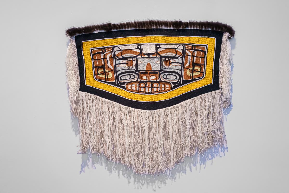 Installation photograph of Lily Hope's Between Worlds child's robe, with a broad cream field, black, blue, and brown curving forms, a yellow border, and long wool fringe.