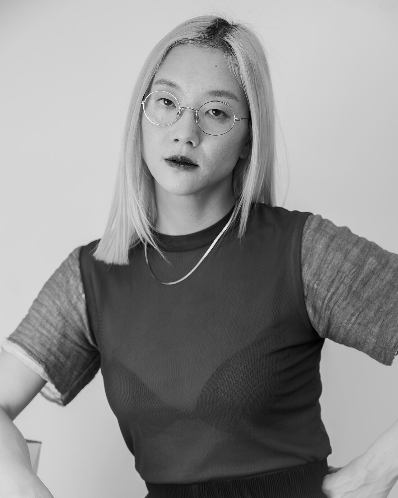 Black-and-white portrait of Christine Sun Kim facing the camera in round glasses and layered dark clothing against a pale wall.