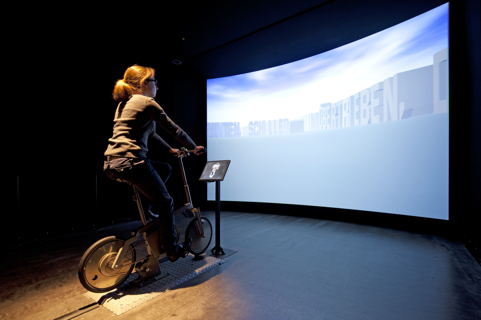 Installation photograph of a participant riding the stationary bicycle in The Legible City before a curved projection of pale three-dimensional letters arranged as streets.
