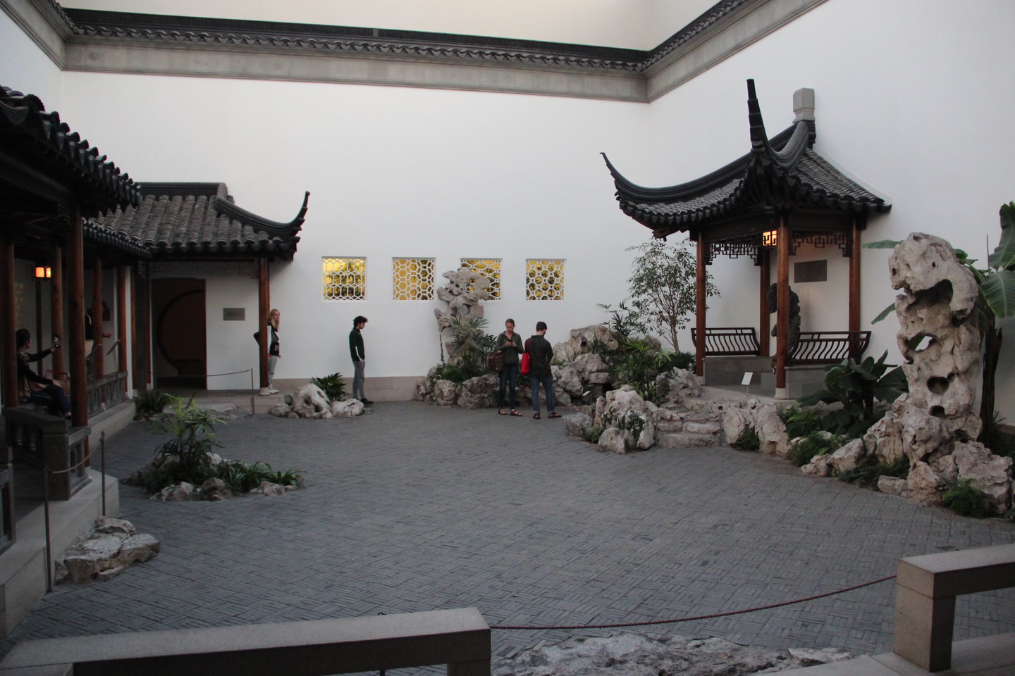 Wide photographic view of The Met's Astor Chinese Garden Court, with dark tiled roofs, timber pavilions, perforated Taihu rocks, plants, and patterned gray paving enclosed by white museum walls.