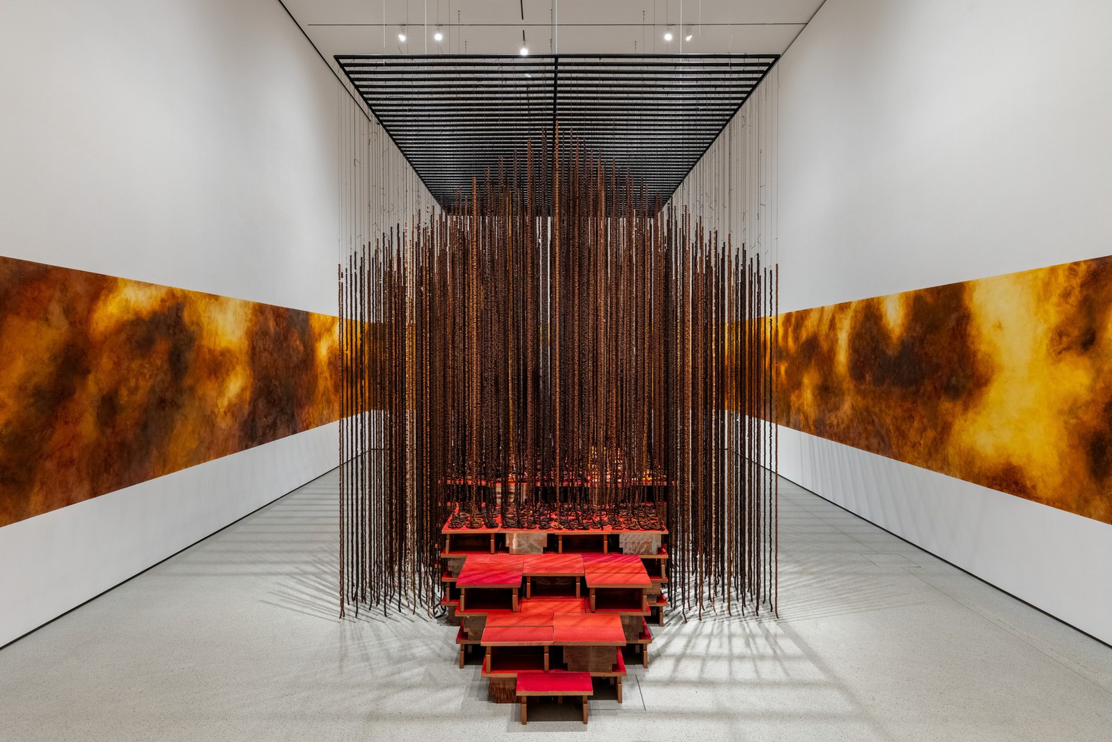 Installation view of Montien Boonma's House of Hope, with a dense house-shaped curtain of dark herbal beads above tiered red stools and ochre cloud-like murals extending along both gallery walls.