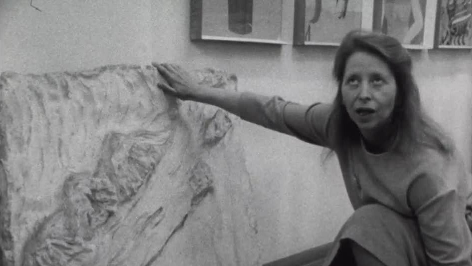 Black-and-white archival film still of sculptor Rolanda Polonsky kneeling beside a large textured plaster relief, with one hand resting along its upper edge.