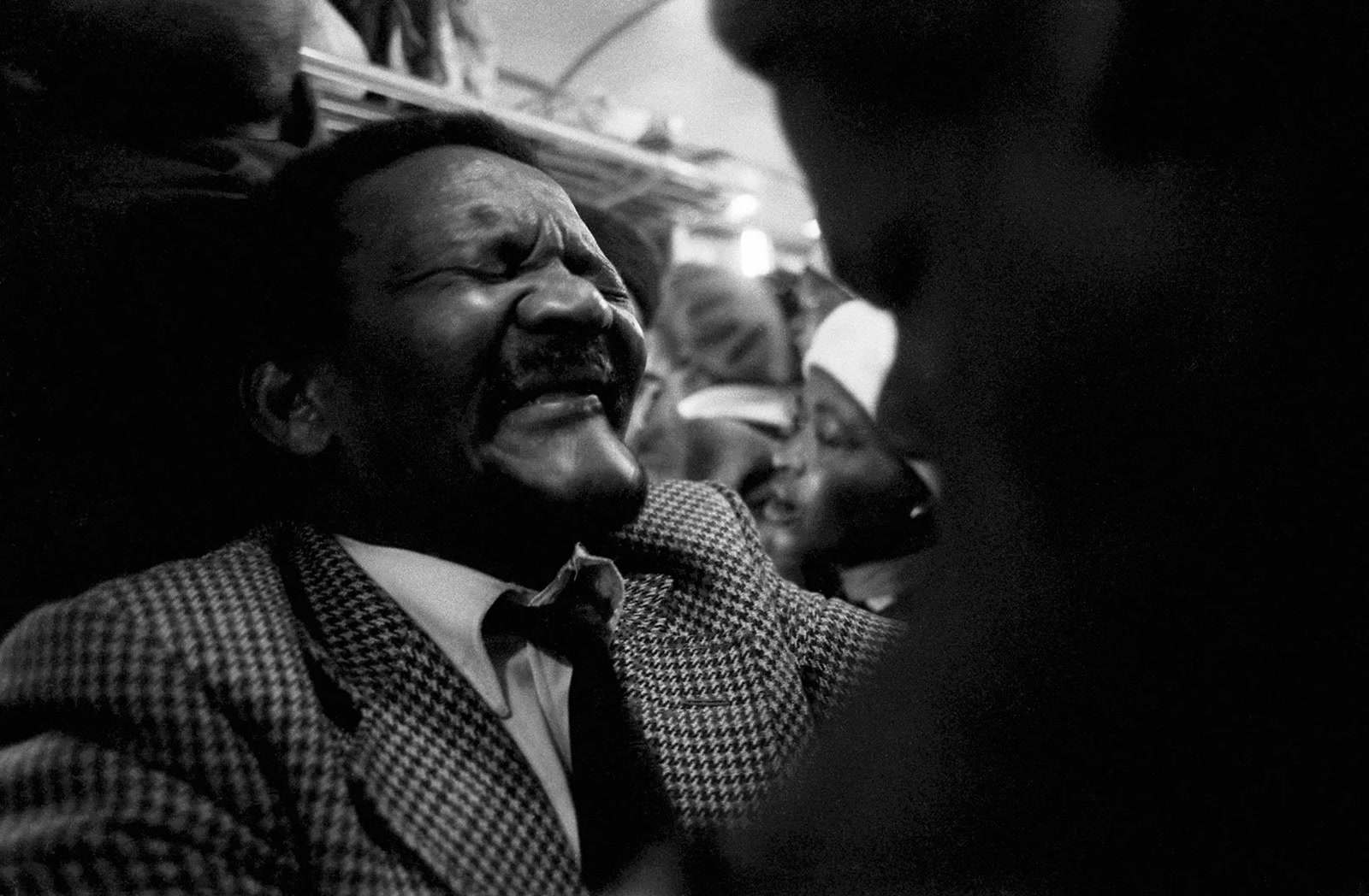 Black-and-white close-up of a worshipper in a checked jacket with eyes shut and mouth open during a crowded church service on a Johannesburg-Soweto commuter train.