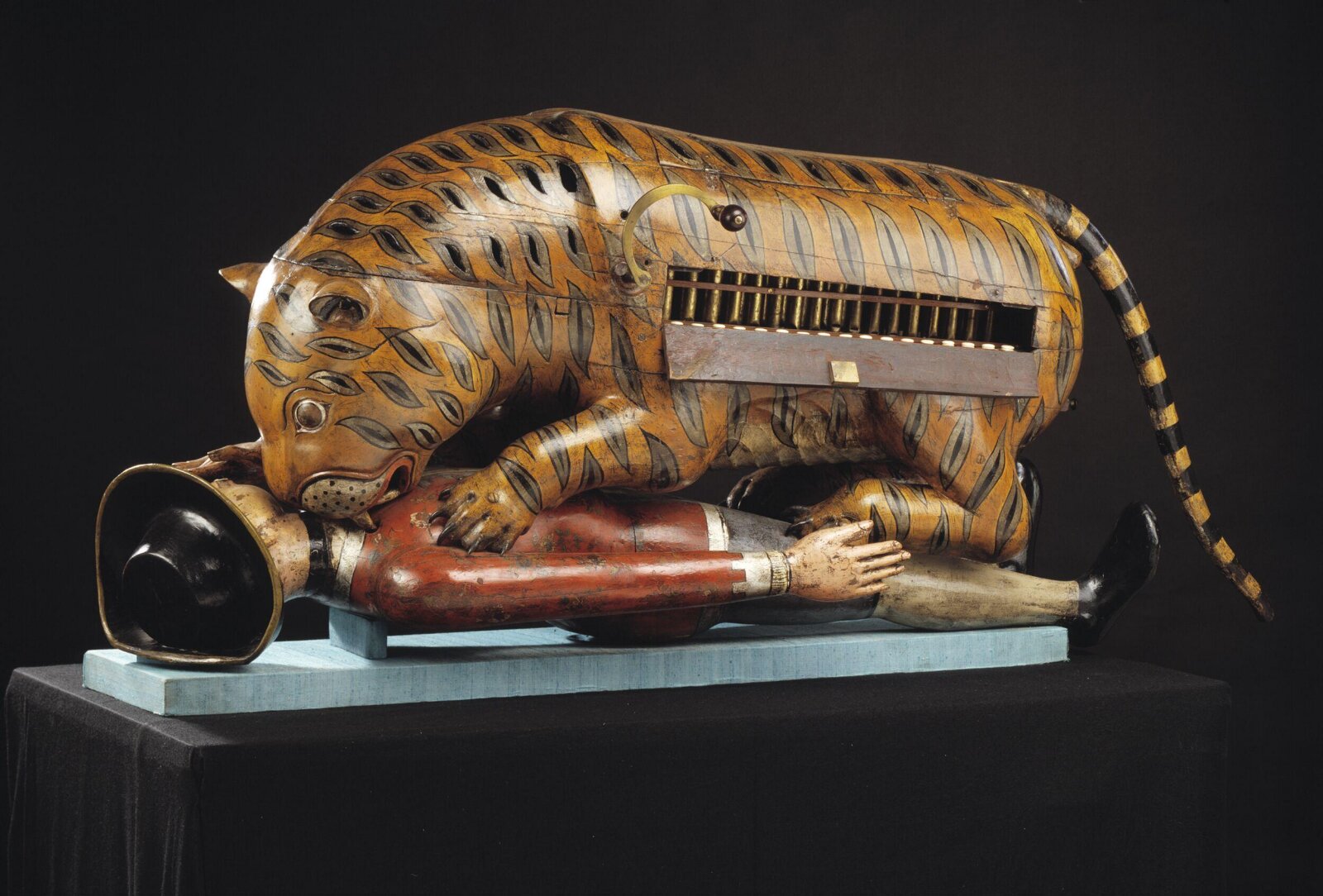 Painted wooden tiger crouching over a life-size European figure, with a hinged panel open on its flank to reveal organ pipes and button keys.