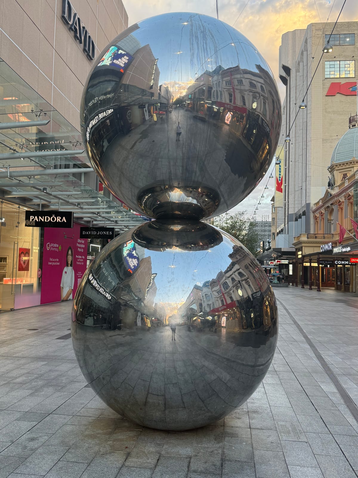 两枚上下叠放的抛光不锈钢球立在 Rundle Mall，映出步行街、黄昏中的阿德莱德店面，以及独自站在其间的摄影者。