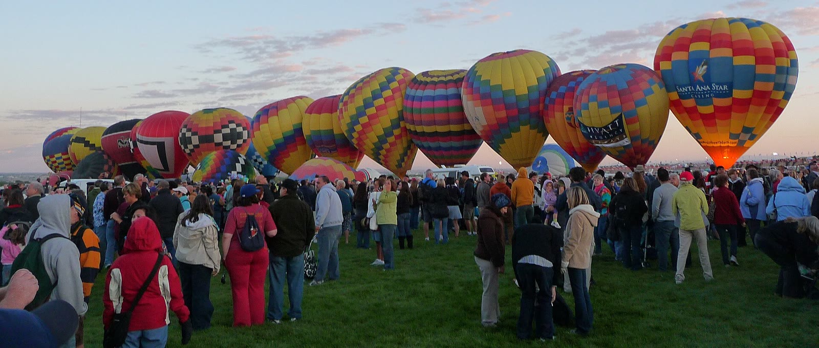 Spectators in jackets stand among rows of colorful hot-air balloons inflating across the Albuquerque Balloon Fiesta launch field at dawn.