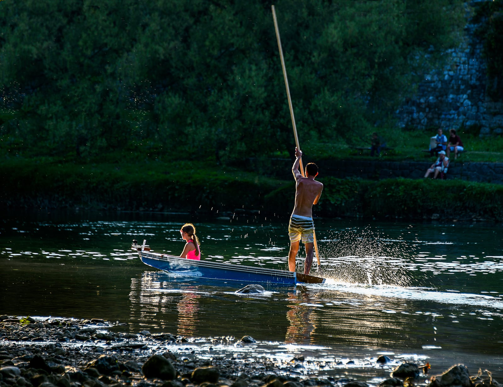 A standing boatman drives a long pole into the Vrbas while a passenger sits low in a narrow blue dajak boat.