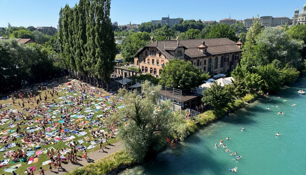 Swimmers float in the turquoise Aare beside a crowded Marzili lawn and the brick Dampfzentrale, with Bern’s skyline above the trees.