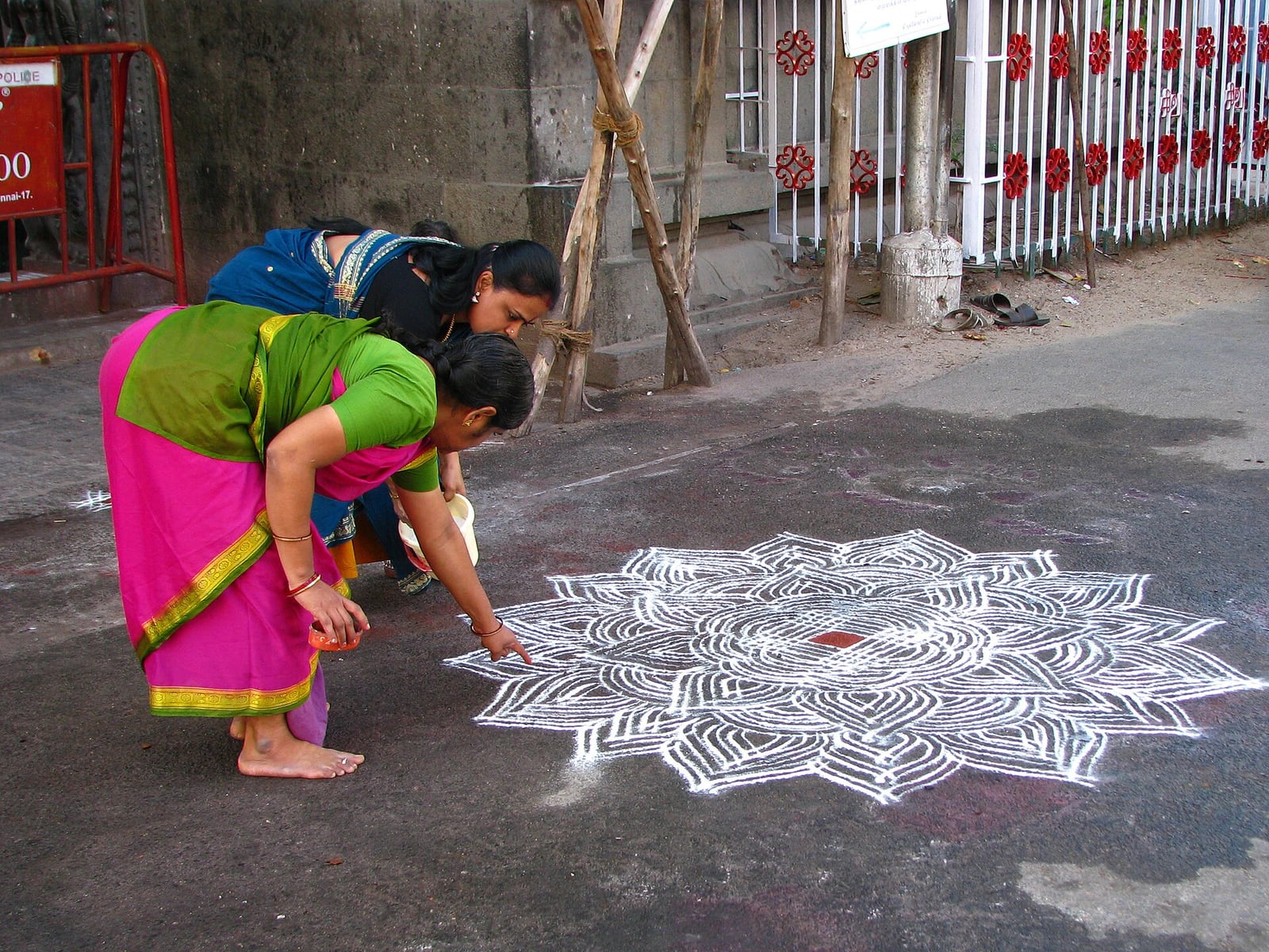 Two women lean over an intricate white kolam on the pavement outside Kapaleeshwarar Temple, with one extending the pattern by hand.
