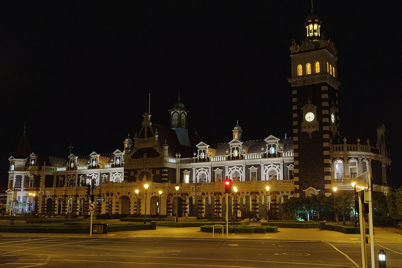 The illuminated dark-stone facade and clock tower of Dunedin Railway Station seen across Anzac Square at night.