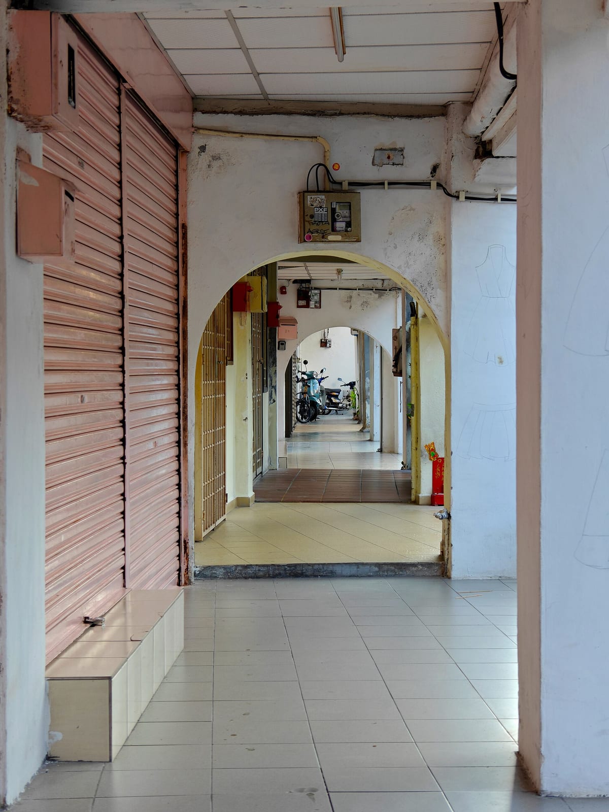A shaded five-foot way recedes beneath a row of George Town shophouses, with tiled thresholds, closed shutters and parked motorcycles.