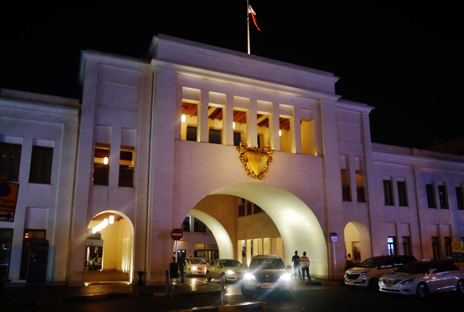 Bab Al Bahrain illuminated at night, with its white central arch framing people and vehicles moving between Customs Square and Manama Souq.
