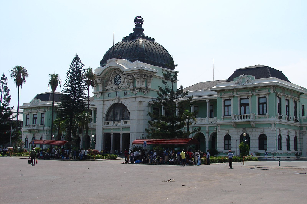 Maputo Central Railway Station’s pale green facade and dark ribbed central dome face Praça dos Trabalhadores beneath a blue sky.