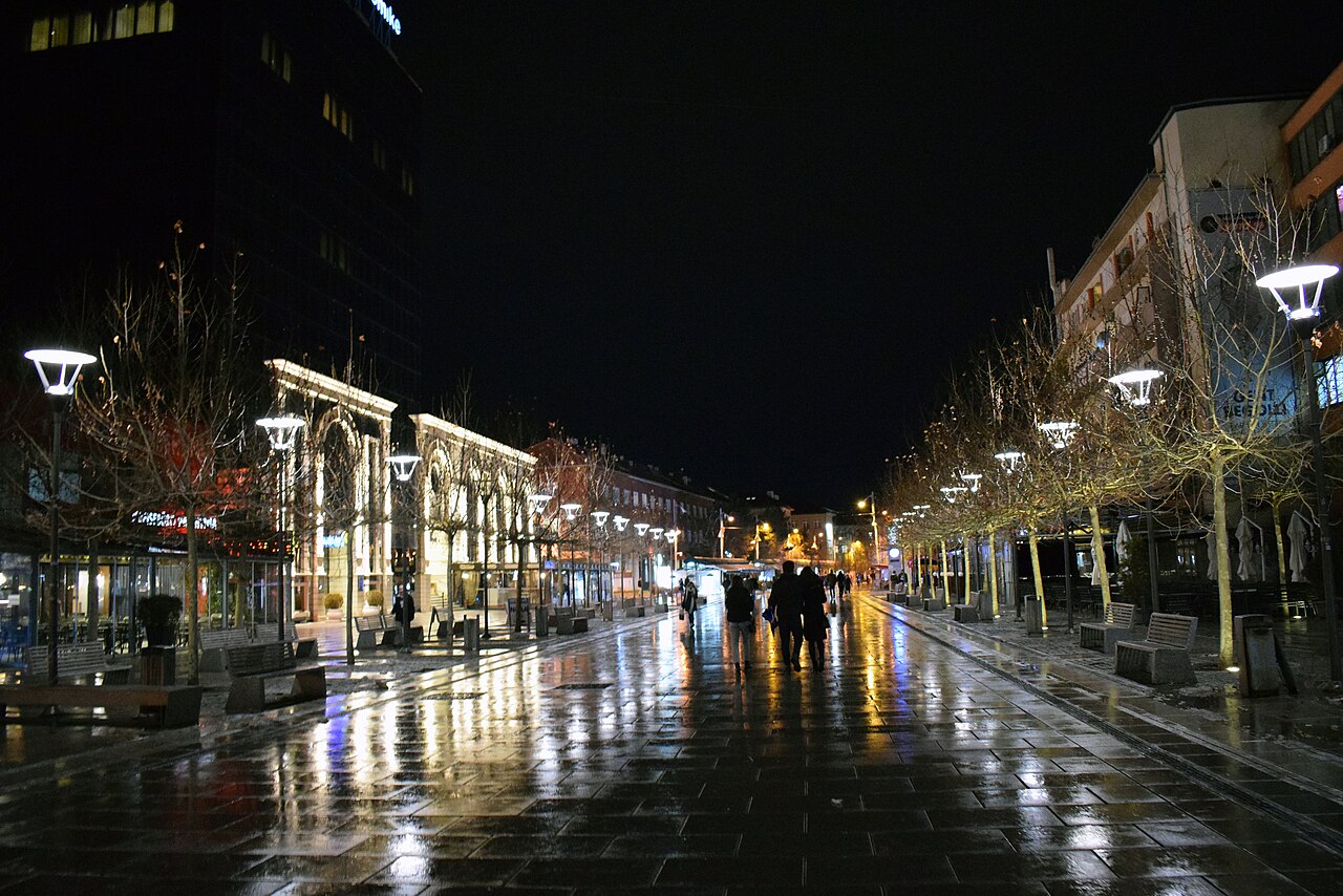 People cross Mother Teresa Square in central Prishtina at night beneath streetlights and illuminated building fronts.