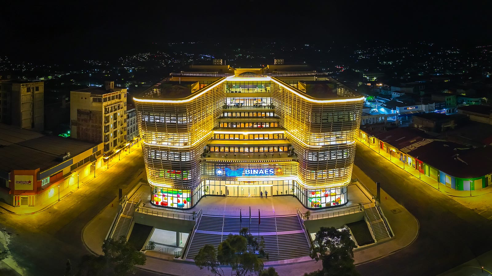 圣萨尔瓦多国家图书馆灯火通明的航拍夜景，可见层叠的玻璃立面、BINAES 标志、宽阔的入口台阶与四周街道。