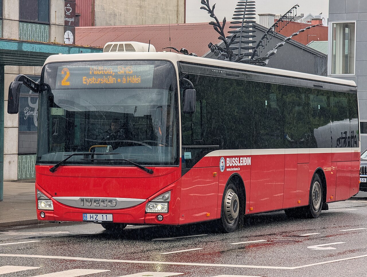一辆红白相间的 Bussleiðin 市内公交行驶在托尔斯港中心被雨水打湿的街道上，车头以法罗语显示 2 路。