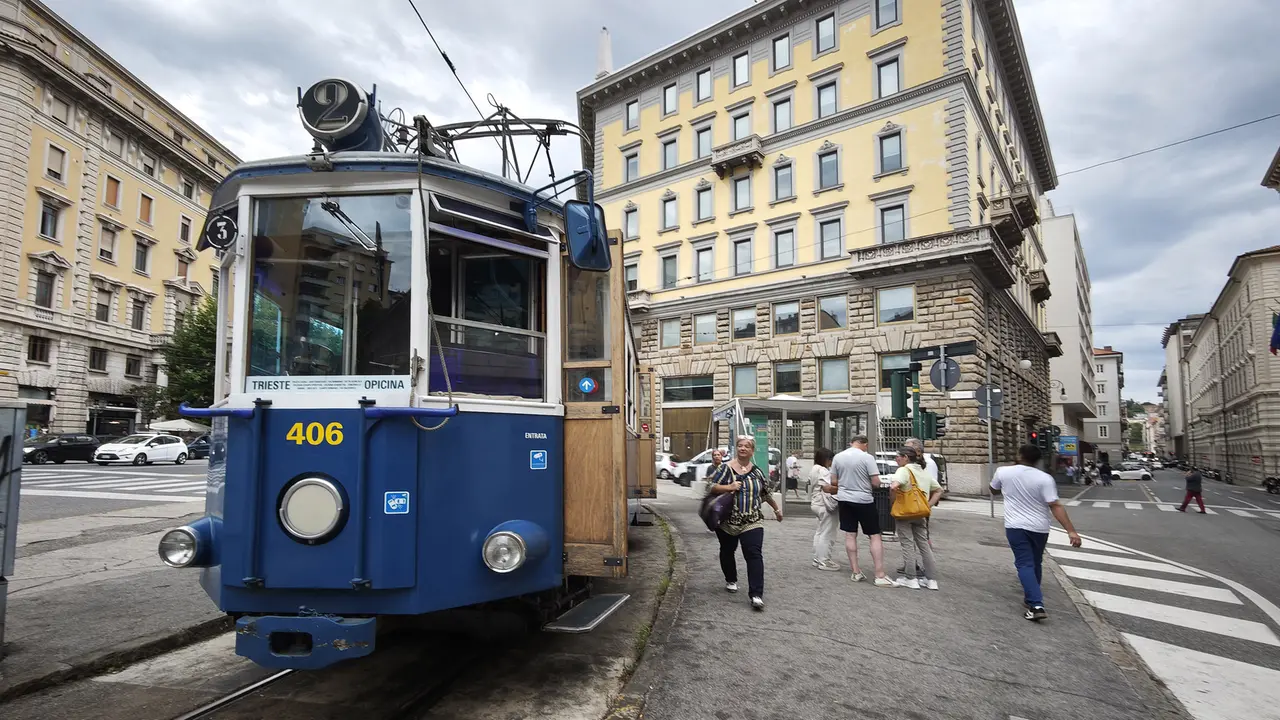 一辆蓝色 Tram di Opicina 电车停在 Piazza Dalmazia 的街面轨道上，行人、架空线与的里雅斯特石砌立面的城市建筑围在四周。