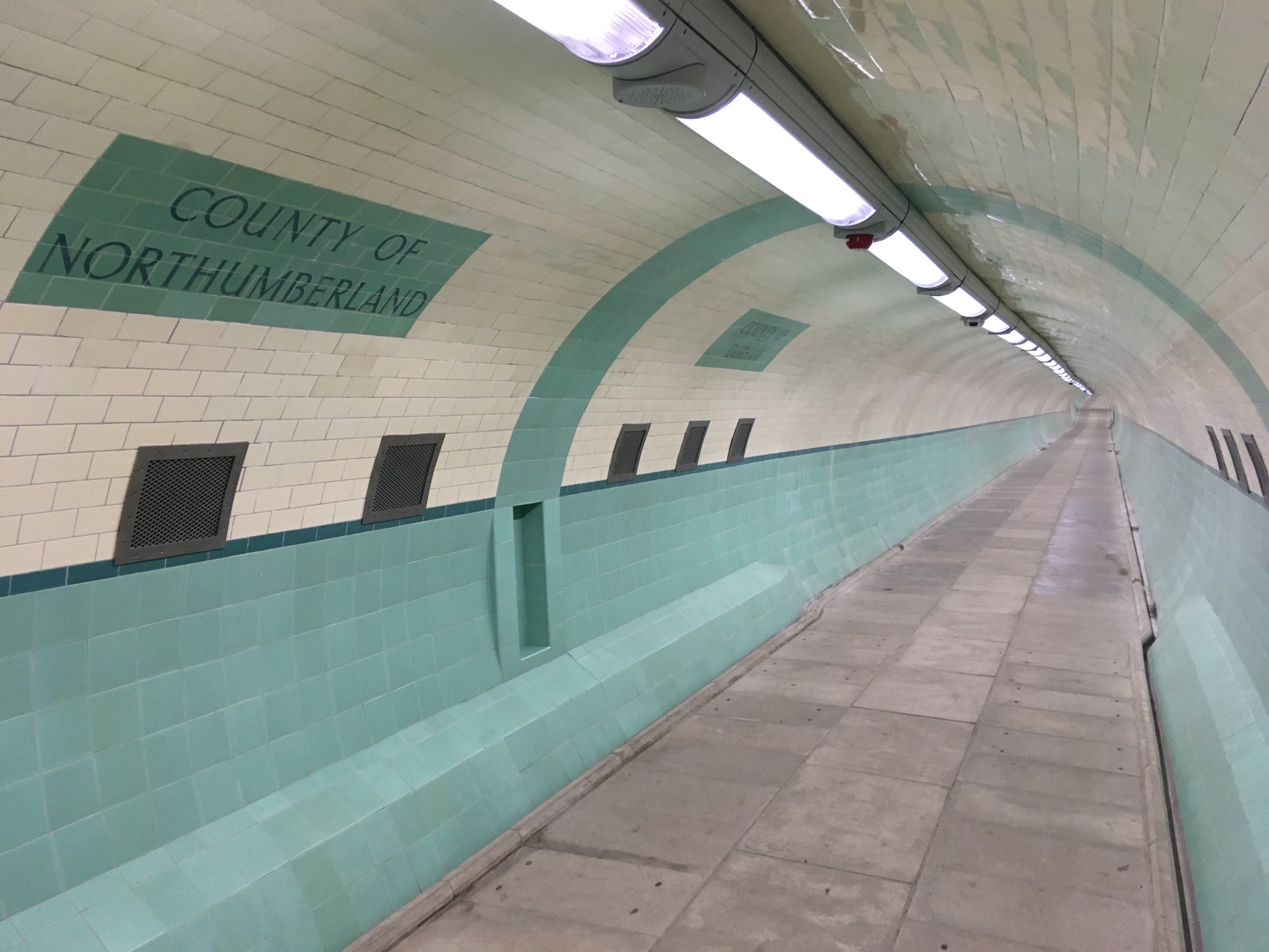 Cream-and-green tiled signs for County Durham and County Northumberland meeting at the subterranean boundary inside the Tyne pedestrian tunnel.