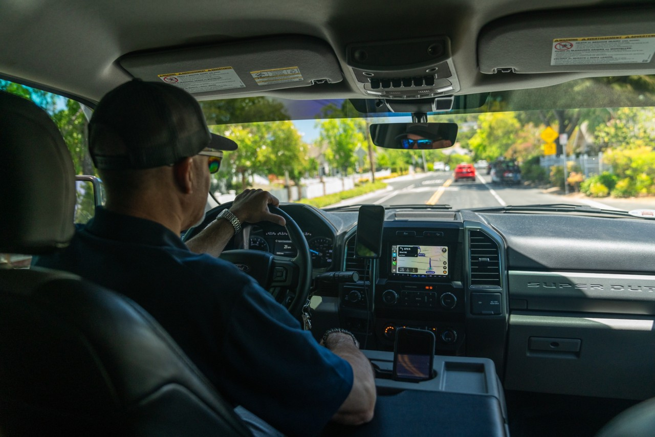 A pool-service professional driving a service truck along a tree-lined residential road.