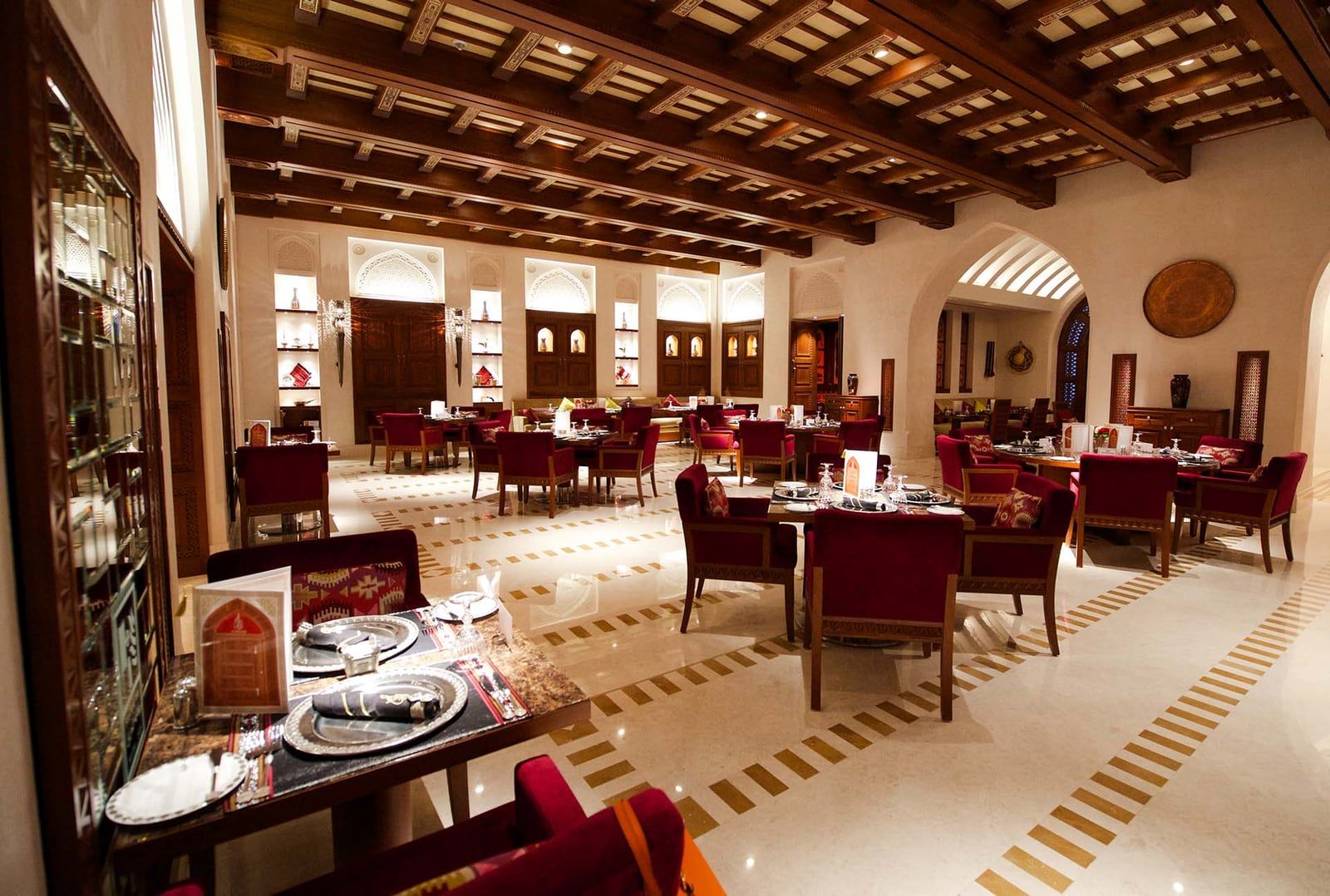 Al Angham's main dining room in Muscat, with set tables, deep-red chairs, carved dark-wood ceiling beams and illuminated Omani arches.
