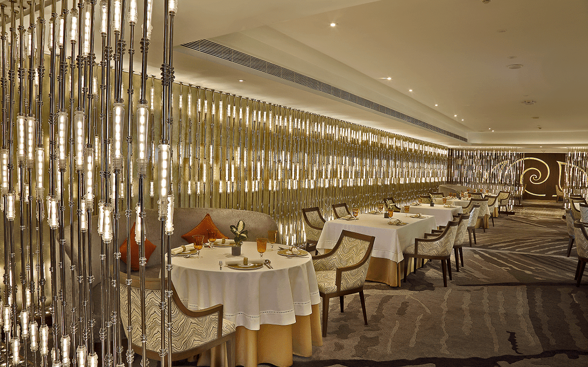 Avartana's long dining room at ITC Grand Chola in Chennai, with white-clothed tables beside rows of warm illuminated glass columns.