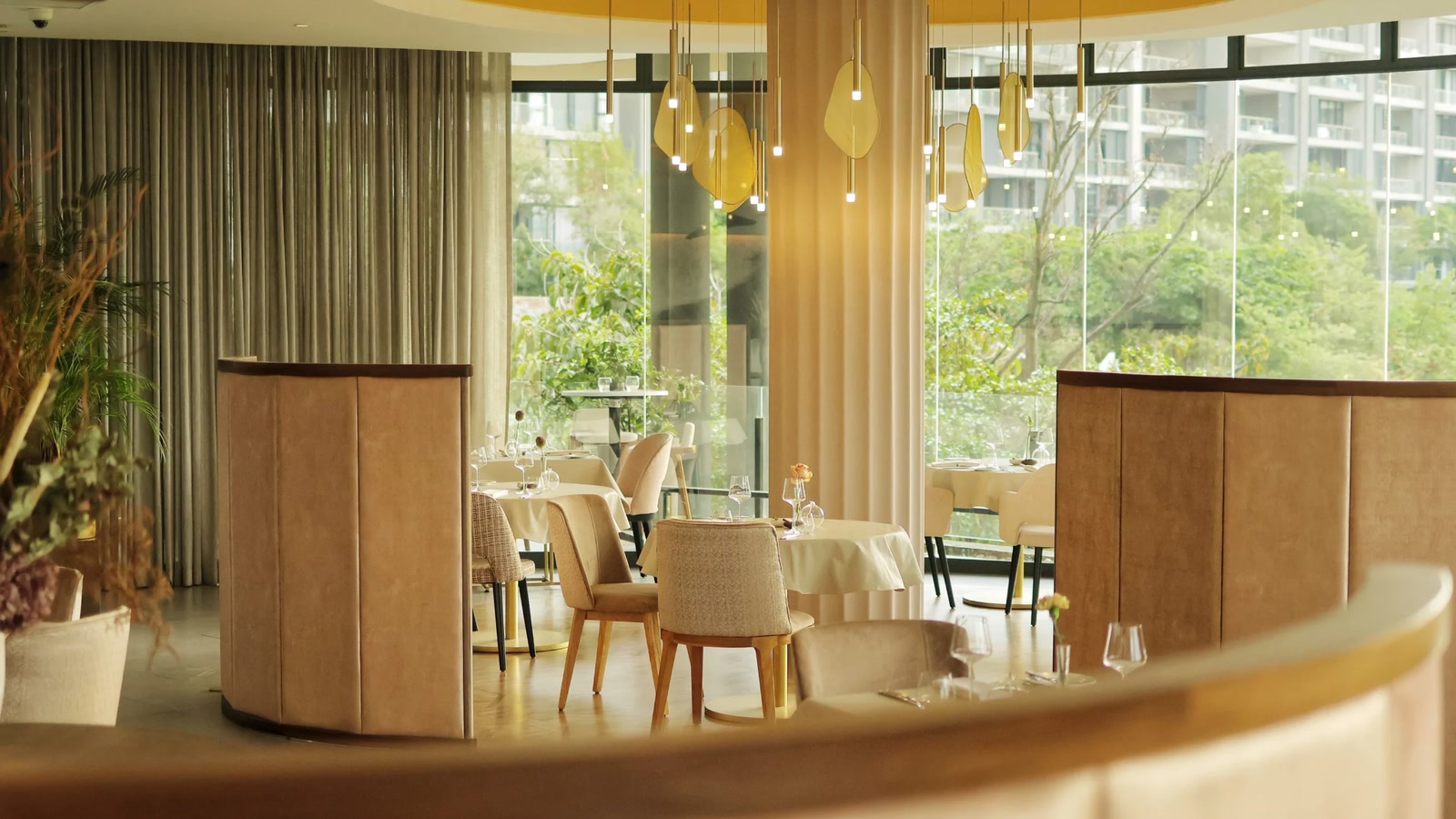 Cyra's dining room in Johannesburg with curved peach banquettes, white-clothed tables, hanging gold lights and greenery visible through tall windows.