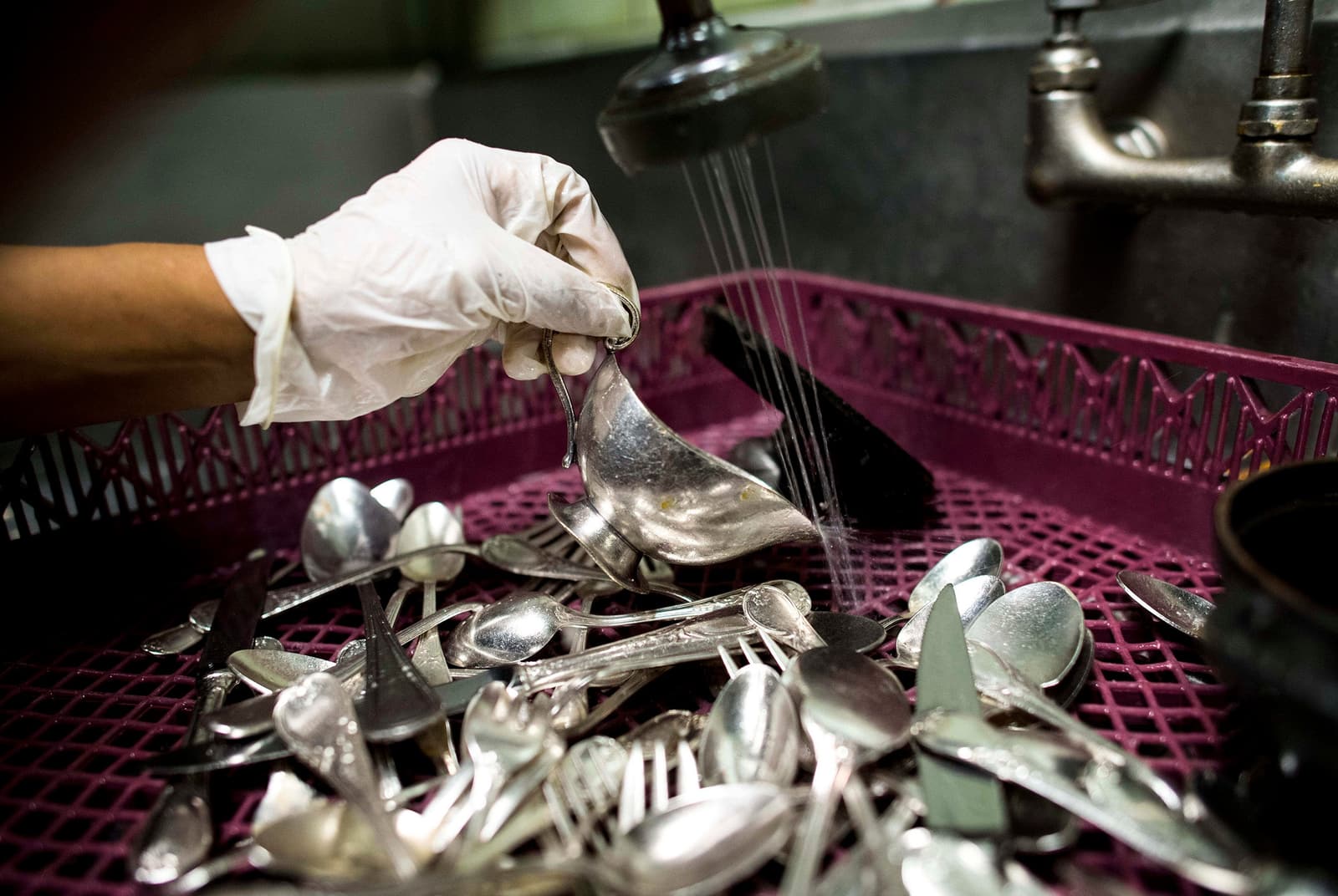 A gloved restaurant worker rinses silver gravy boats, spoons, and flatware in a commercial dishwashing rack.
